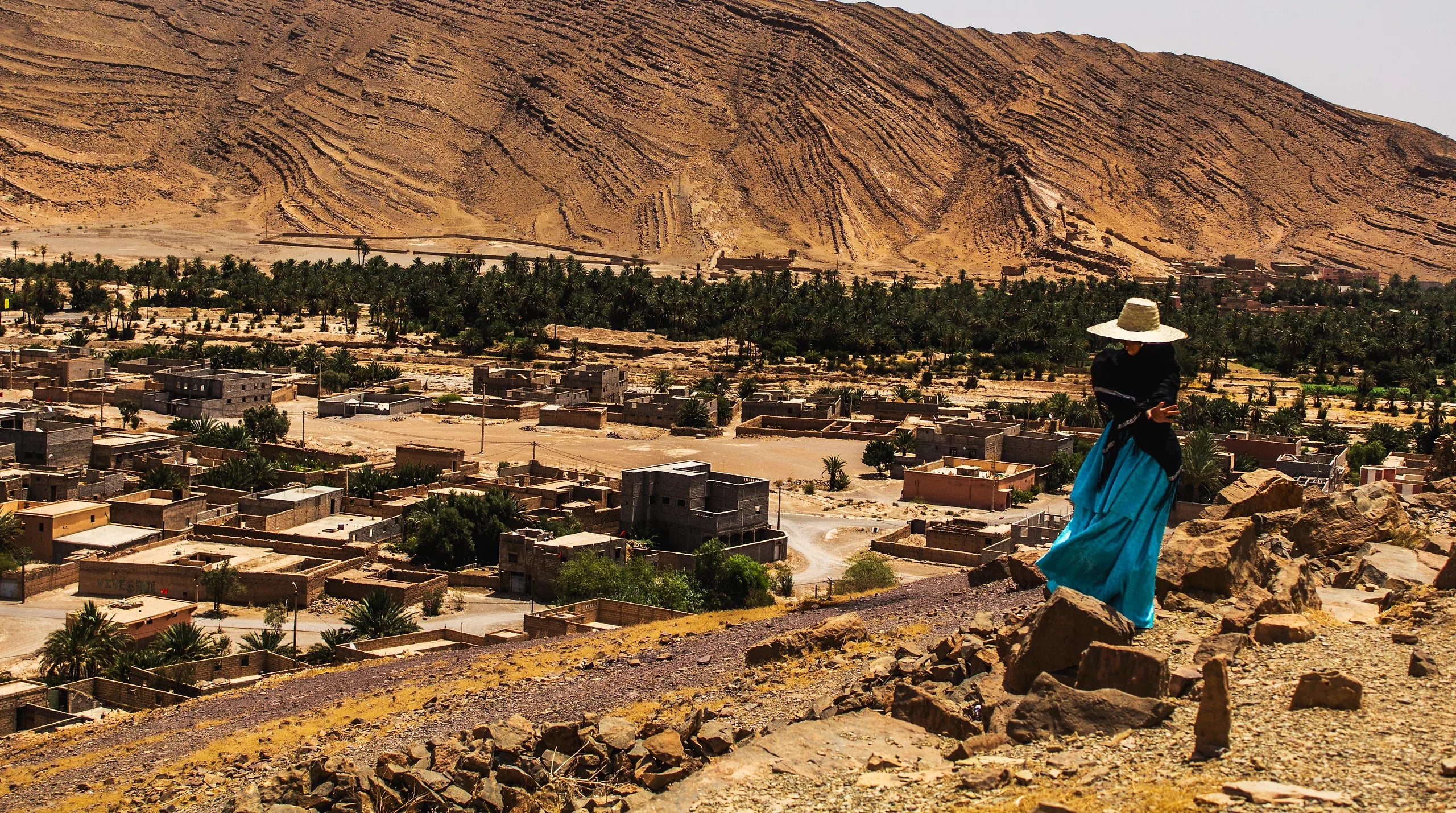 A traveller in a long blue skirt and wide-brimmed straw hat on a rocky path above a palm oasis and village in southern Morocco