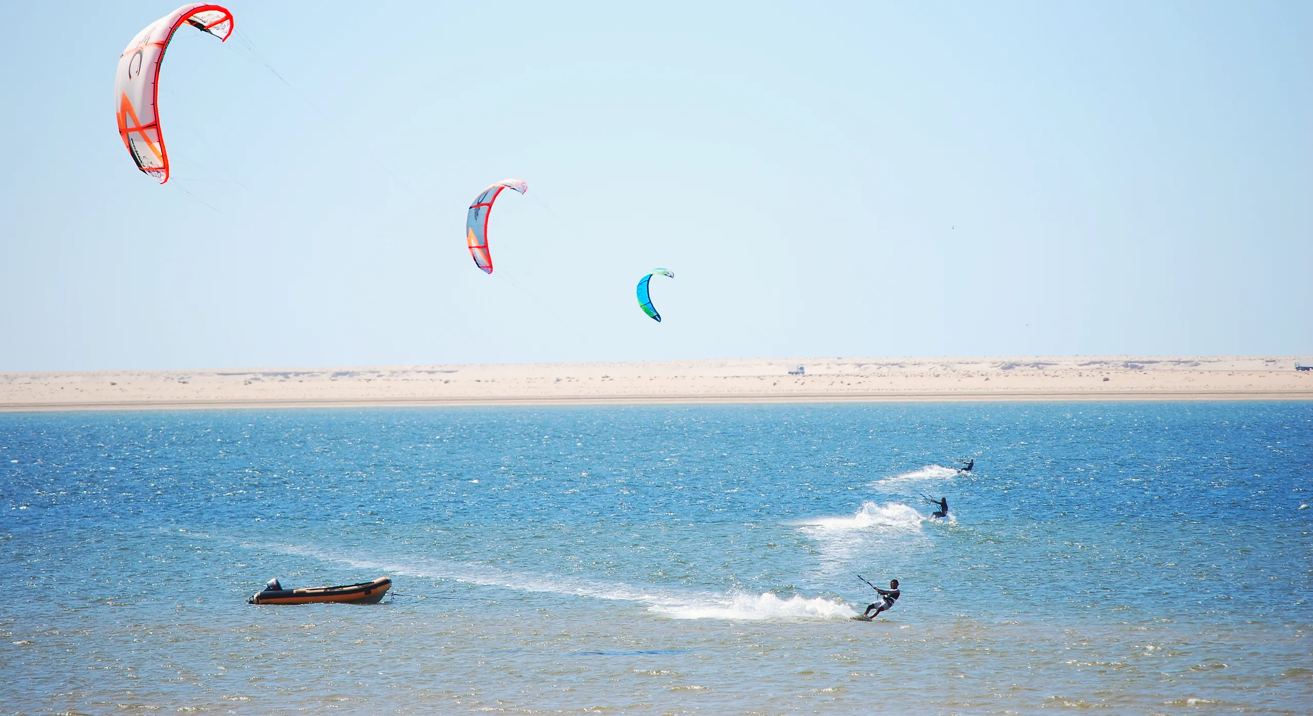 Kitesurfers and a dinghy on the blue Dakhla lagoon below a pale sand shore