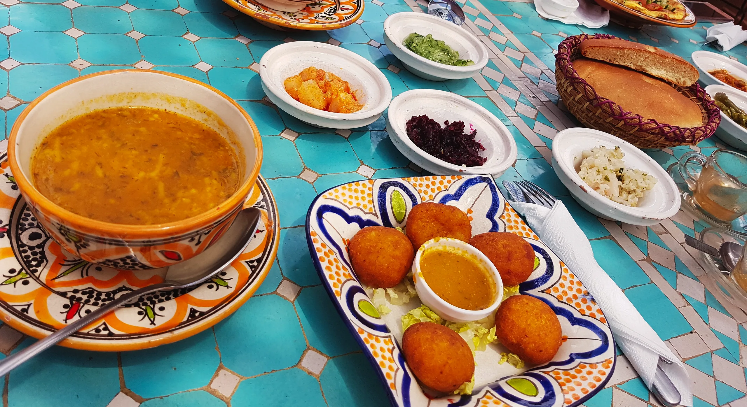 A bowl of harira on a zellige table with fried croquettes, small salads and a basket of bread