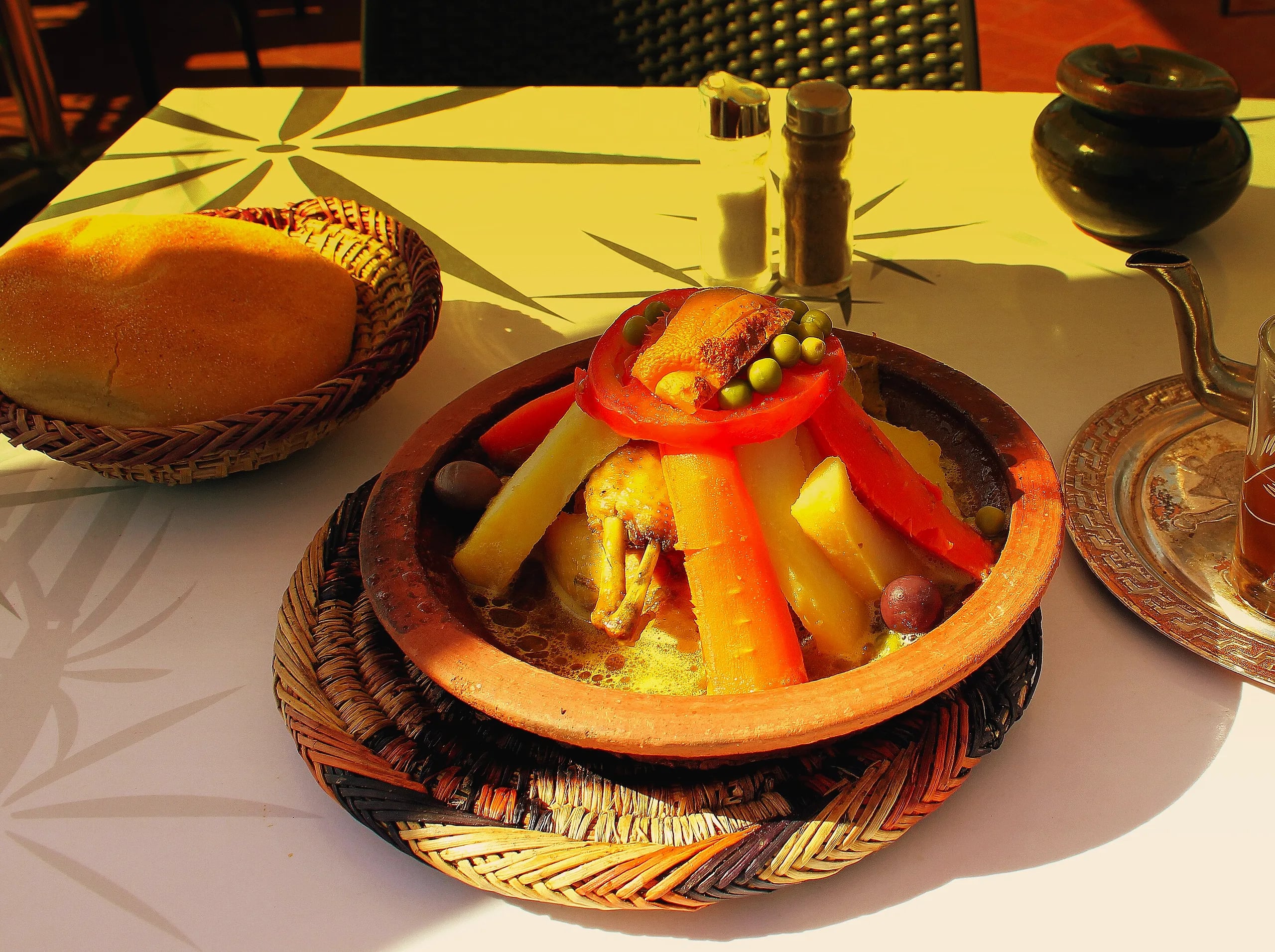 A chicken and vegetable tagine in a clay dish on a woven mat, with a basket of round Moroccan bread and a silver teapot on a café table in Marrakech
